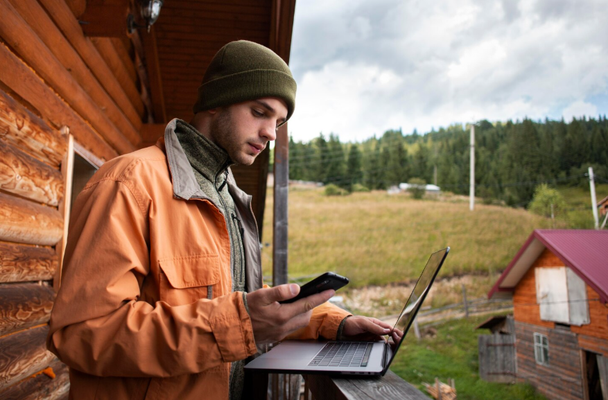 Man using rural internet for streaming with a laptop and phone outside a countryside cabin.